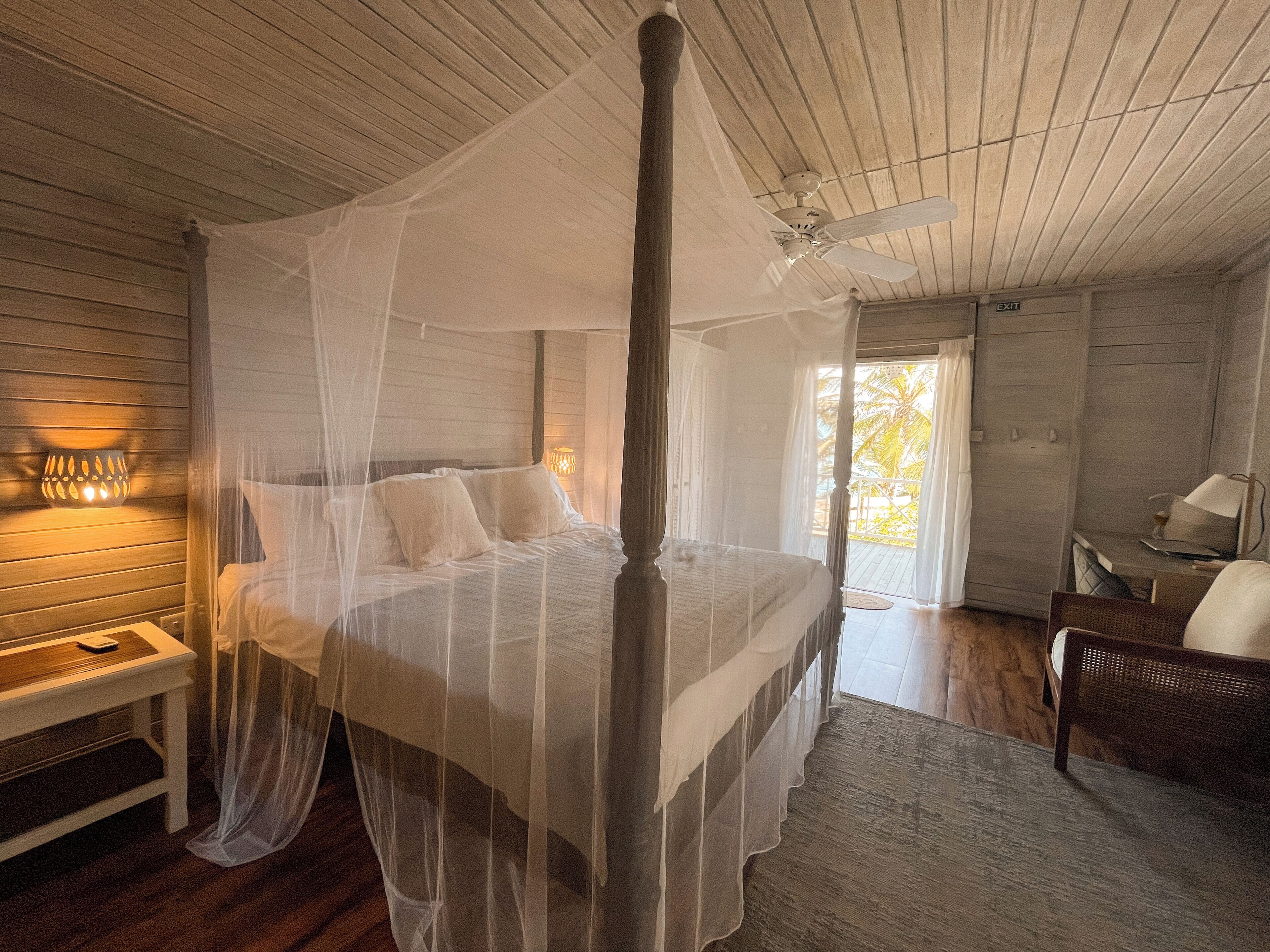 A four-poster bed with a canopy, wooden walls, a bench, a desk, and a chair in a room with a view.