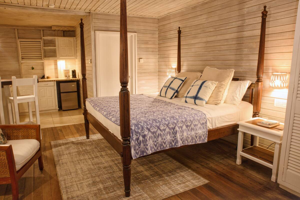 A bedroom with a four-poster bed, a wooden floor, a small bedside table, and a chair.