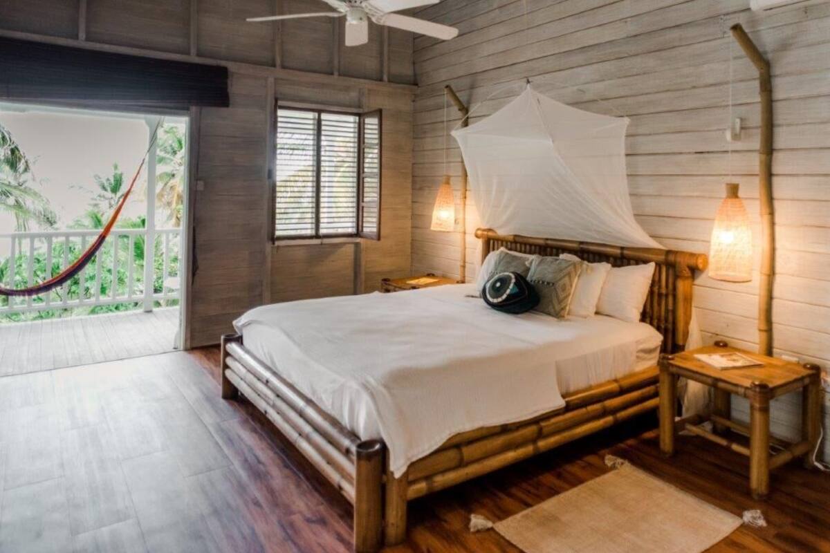 A spacious bedroom with a large bed, wooden walls, a ceiling fan, and a view of greenery through a window.