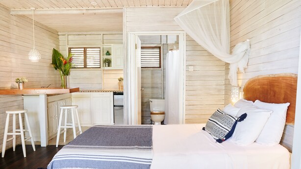 A bedroom with a bed, a bedside table, a nightstand, a window, and a view of the kitchen. - ECO Lifestyle & Lodge (Bathsheba)