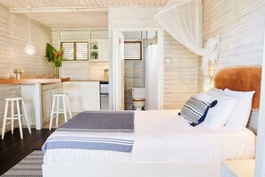 A bedroom with a bed, a bedside table, a nightstand, a window, and a view of the kitchen. - ECO Lifestyle & Lodge (Bathsheba)
