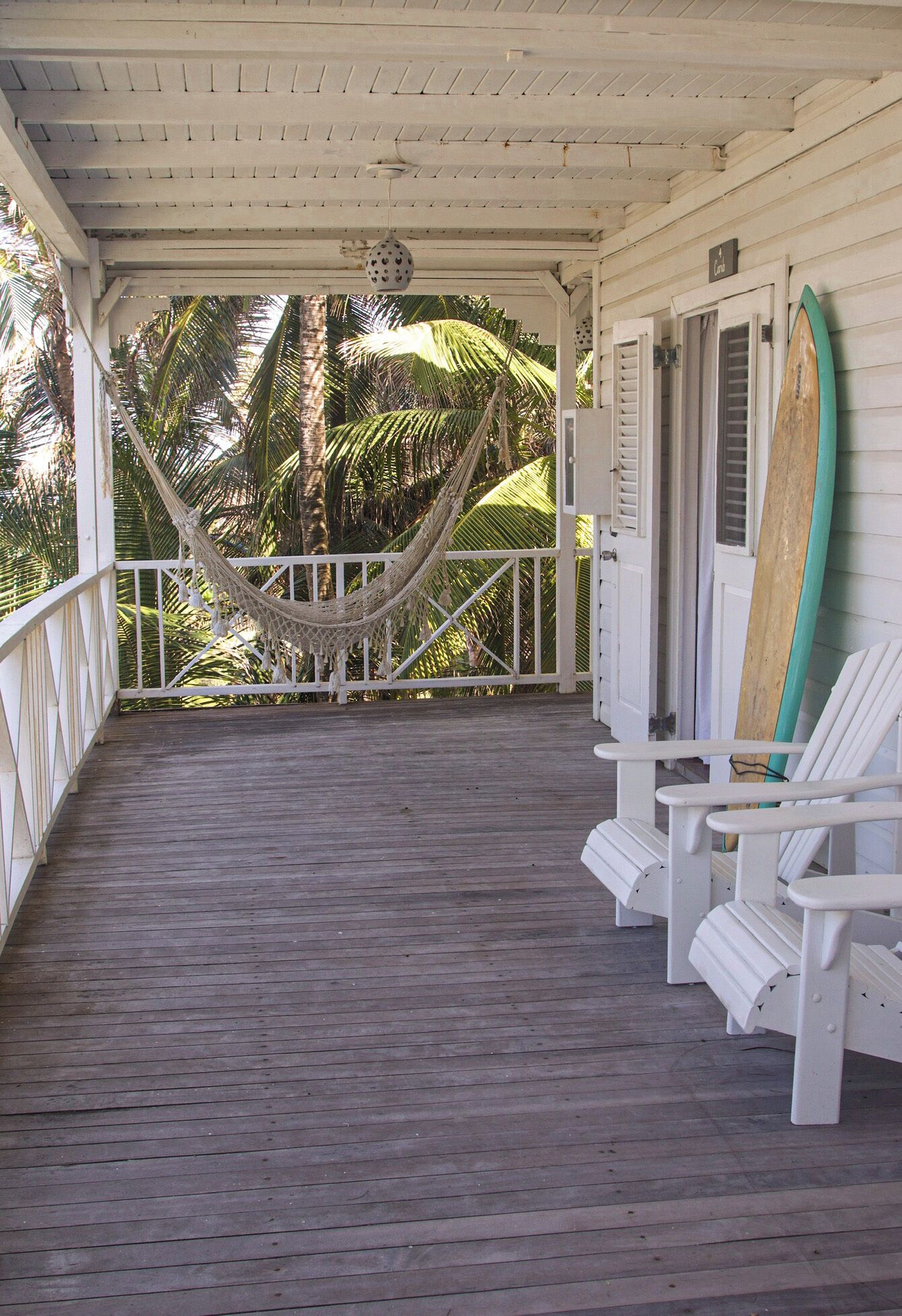 Een houten veranda met een hangmat, witte stoelen en surfplanken tegen de muur.
