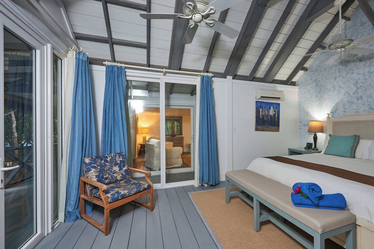 A bedroom with a bed, a bench, a chair, a ceiling fan, and a view through a glass door.