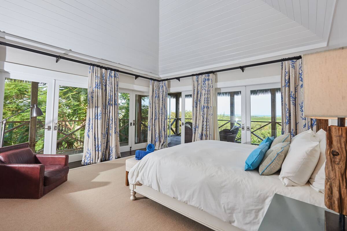 A bedroom with a large bed, a red leather chair, and a view of greenery through the glass doors.