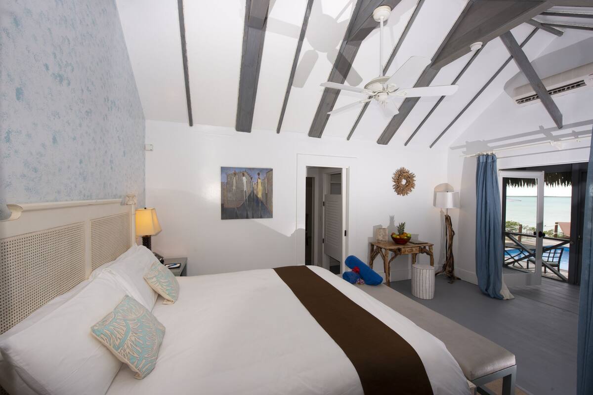 A bedroom with a bed, a ceiling fan, a painting, a desk with a lamp, and a view of the beach.