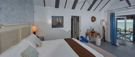 Habitación de hotel con cama, mesita de noche, lámpara, un cuadro, una pequeña mesa con un frutero, lámpara de pie y vista al océano.