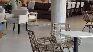 A spacious indoor area with a high ceiling, featuring a dark brown sofa, white armchairs, and wicker tables. - Cara Hotels Trinidad (Claxton Bay)