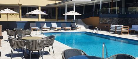 A poolside area with lounge chairs and umbrellas, a clear blue pool, and a building with a covered patio.