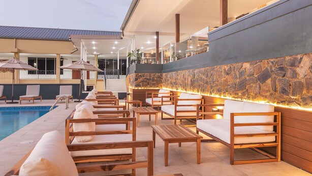A poolside area with wooden lounge chairs and a stone wall with a built-in fire feature. - Cara Hotels Trinidad (Claxton Bay)