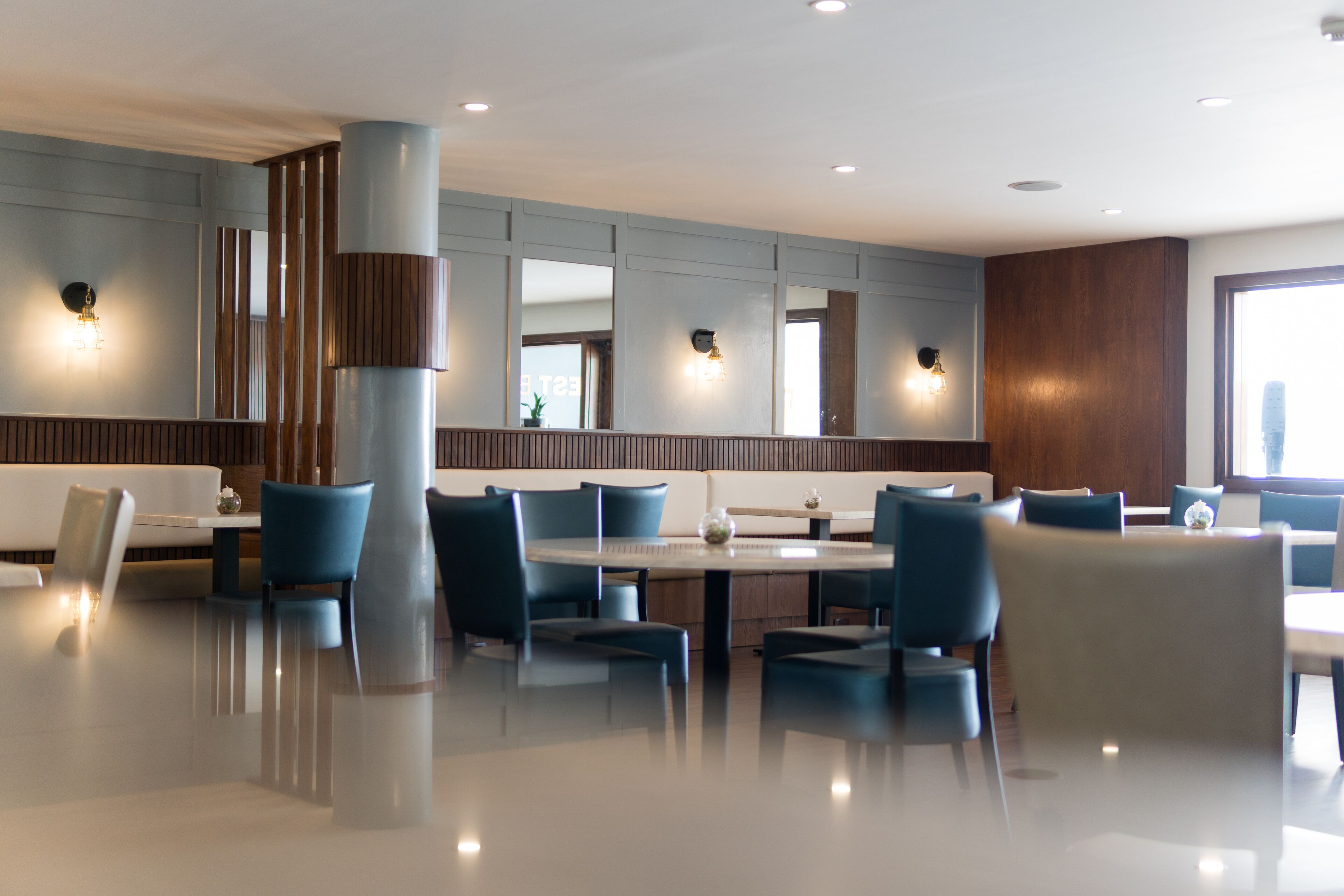 A modern restaurant with a clean, minimalist design, featuring a central dining area with round tables and blue chairs, surrounded by wooden paneling and recessed lighting.