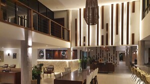 A spacious, modern hotel lobby with a high ceiling, a central island dining table, and a reception desk. - Cara Hotels Trinidad (Claxton Bay)
