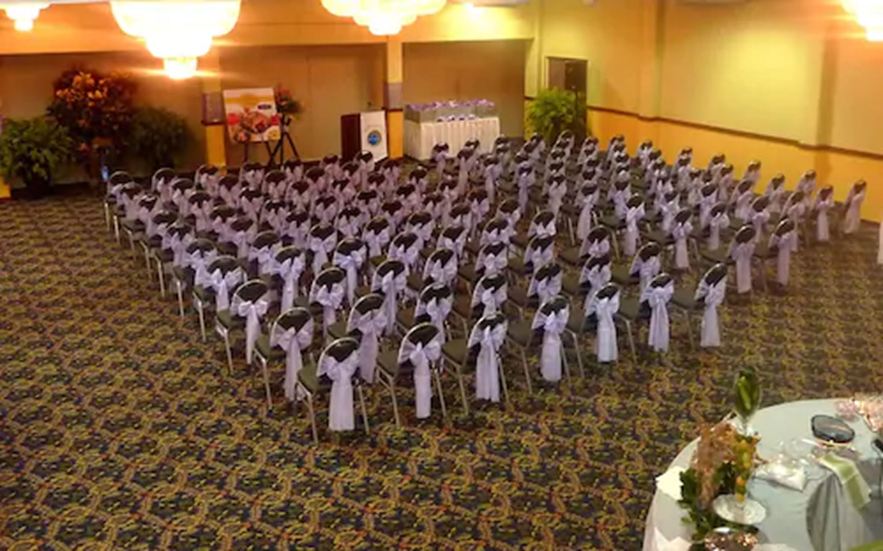 A room with rows of chairs arranged in a semi-circle, a buffet table with a floral centerpiece, and a stage with a speaker and decorations.