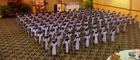 A room with rows of chairs arranged in a semi-circle, a buffet table with a floral centerpiece, and a stage with a speaker and decorations.