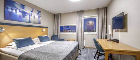 A hotel room with a bed, a desk, a chair, a TV, a window with curtains, and a cityscape view.