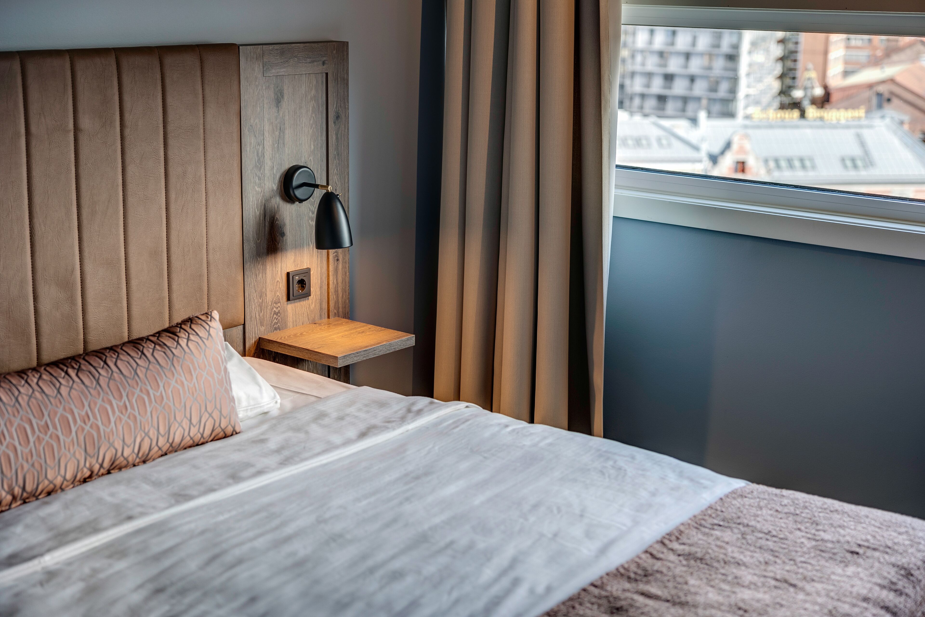A modern hotel room with a wooden headboard, a bedside table with a lamp, and a view of the city through a window.