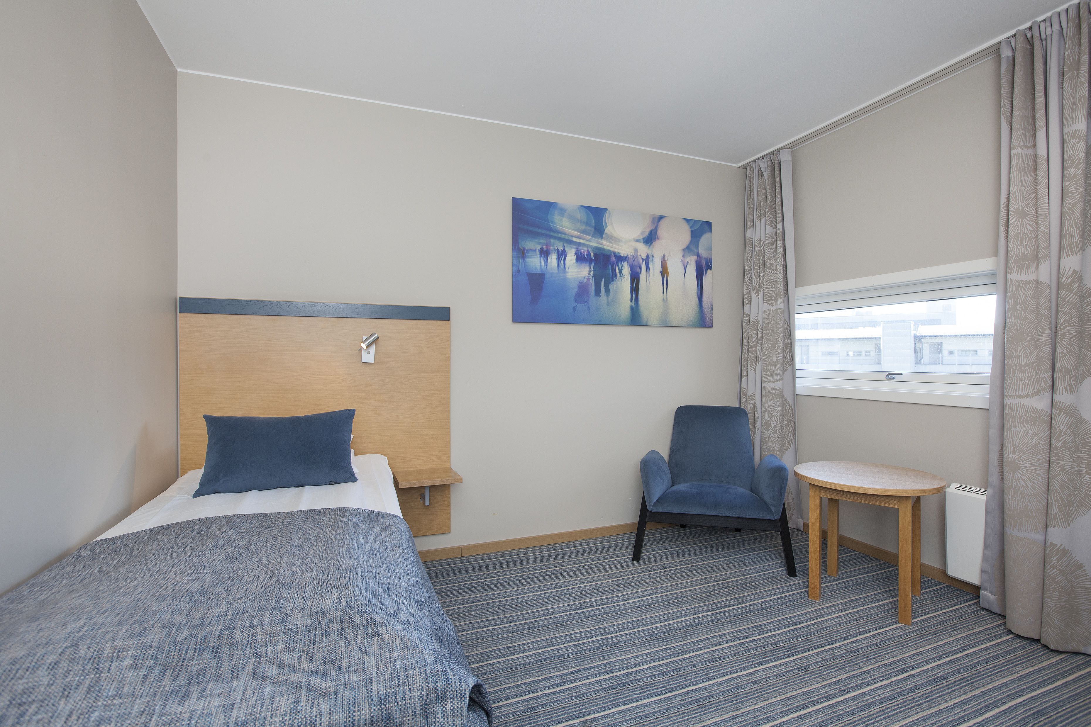 Hotelkamer met een bed, een stoel, een tafeltje, een schilderij aan de muur en een raam met gordijnen.