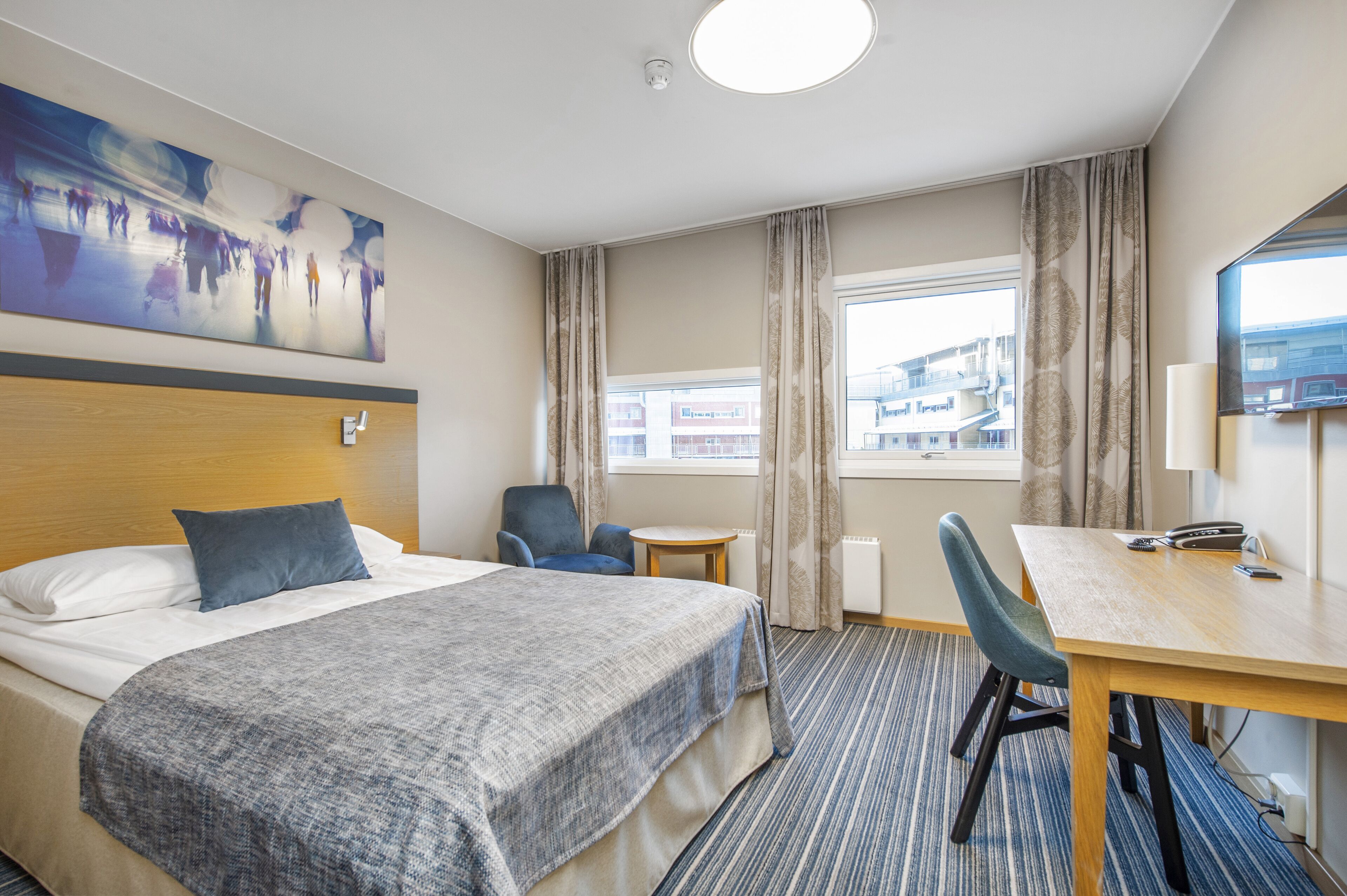 Ein Hotelzimmer mit Bett, Schreibtisch, Stuhl, einem fenster mit Vorhängen, einem Bild an der Wand und Blick auf Gebäude.