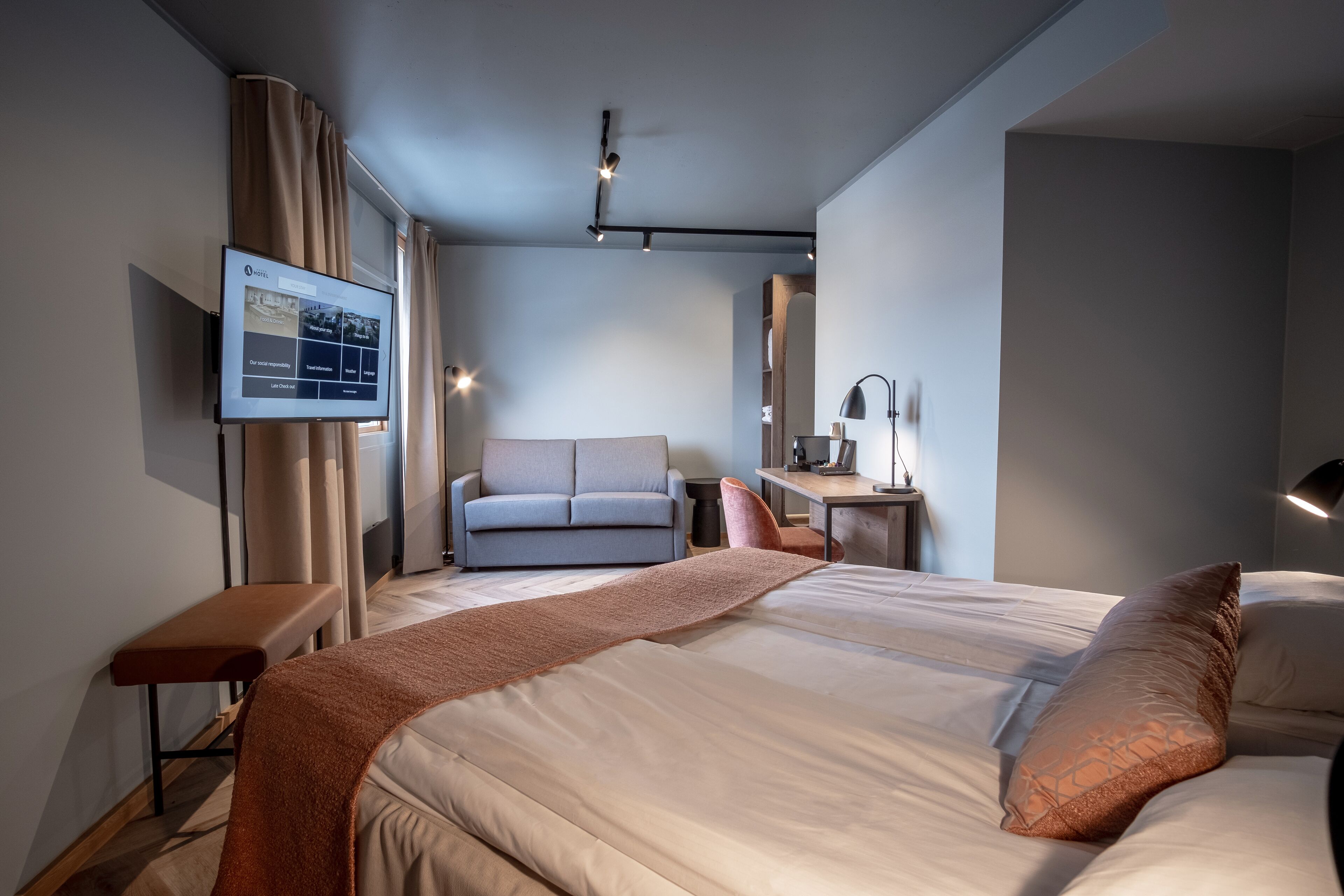 Een moderne hotelkamer met een groot bed, een bureau, een bank en een televisie waarop kamerinformatie wordt weergegeven.