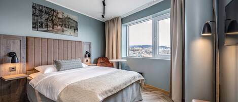 Una habitación de hotel moderna con una cama grande, vista a la ciudad, cabecero de madera y una mesita de noche con lámpara.