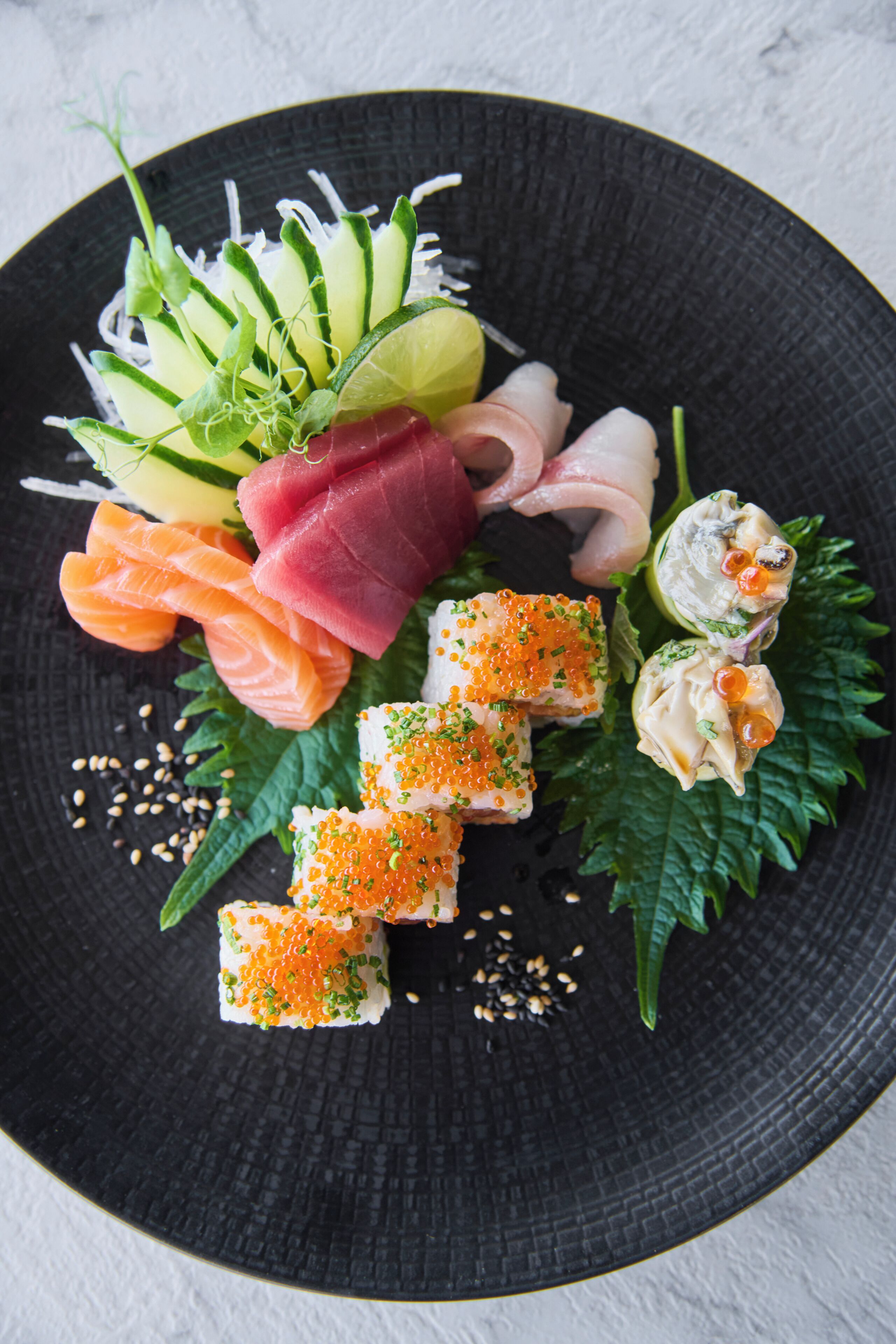 Een zwart bord met verschillende soorten sushi, waaronder sashimi en nigiri, versierd met groene bladeren en sesamzaadjes.