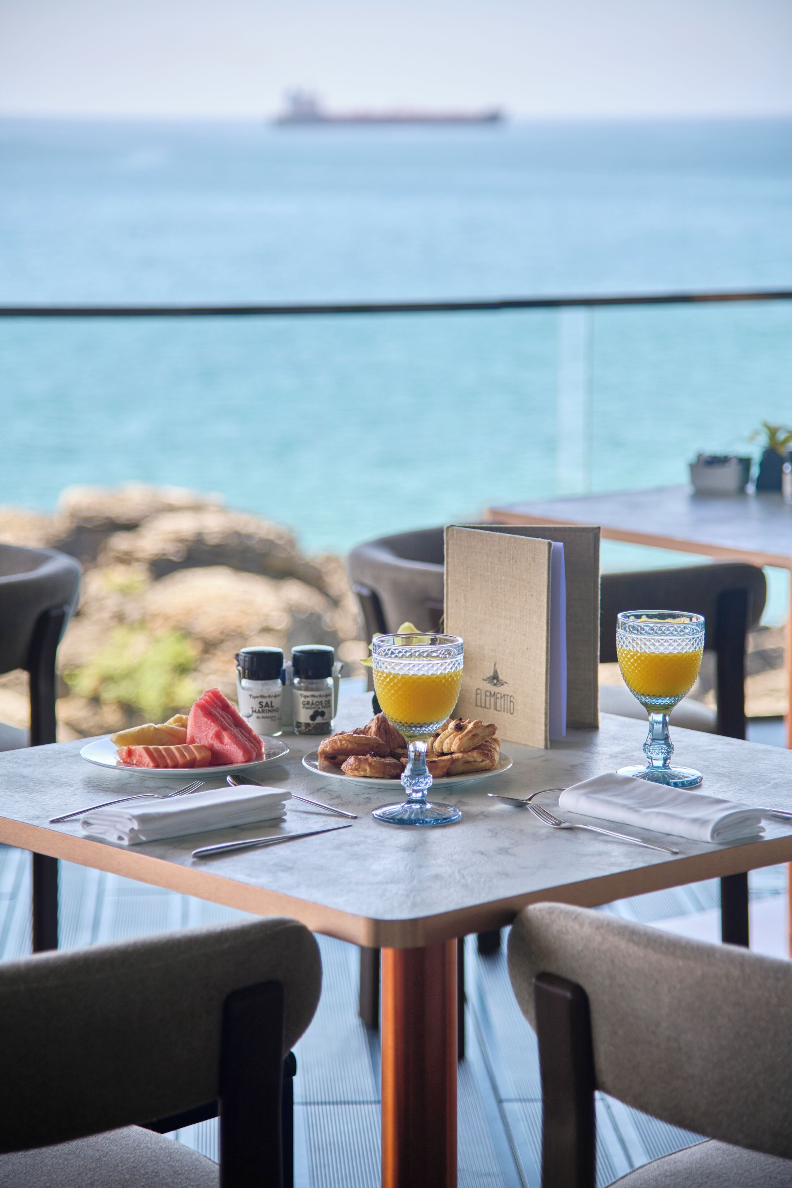 A table set with breakfast items and a glass of orange juice, overlooking a body of water with a ship in the distance.
