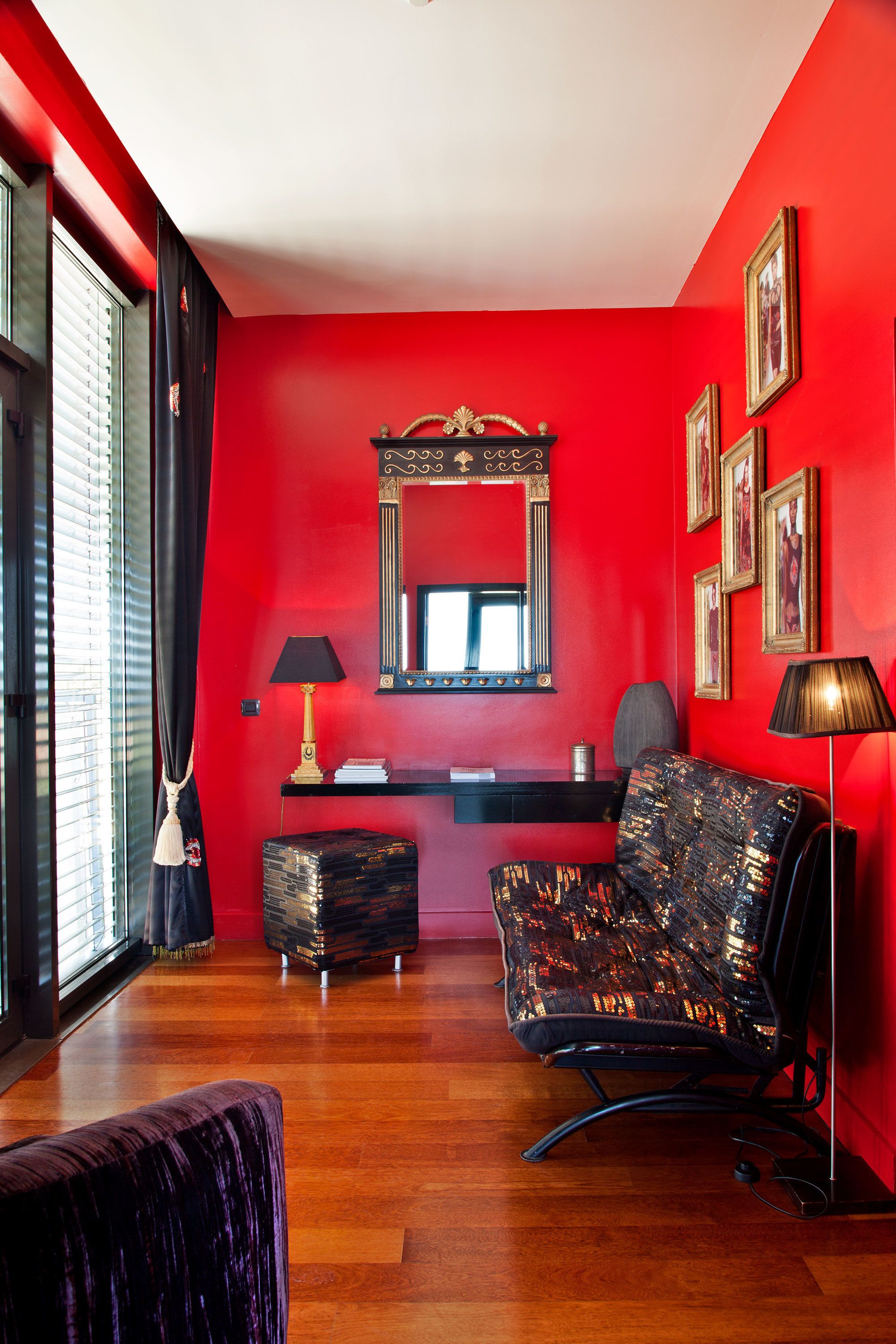 A room with a red wall, a black chair, a black ottoman, a desk with a lamp, and a mirror.