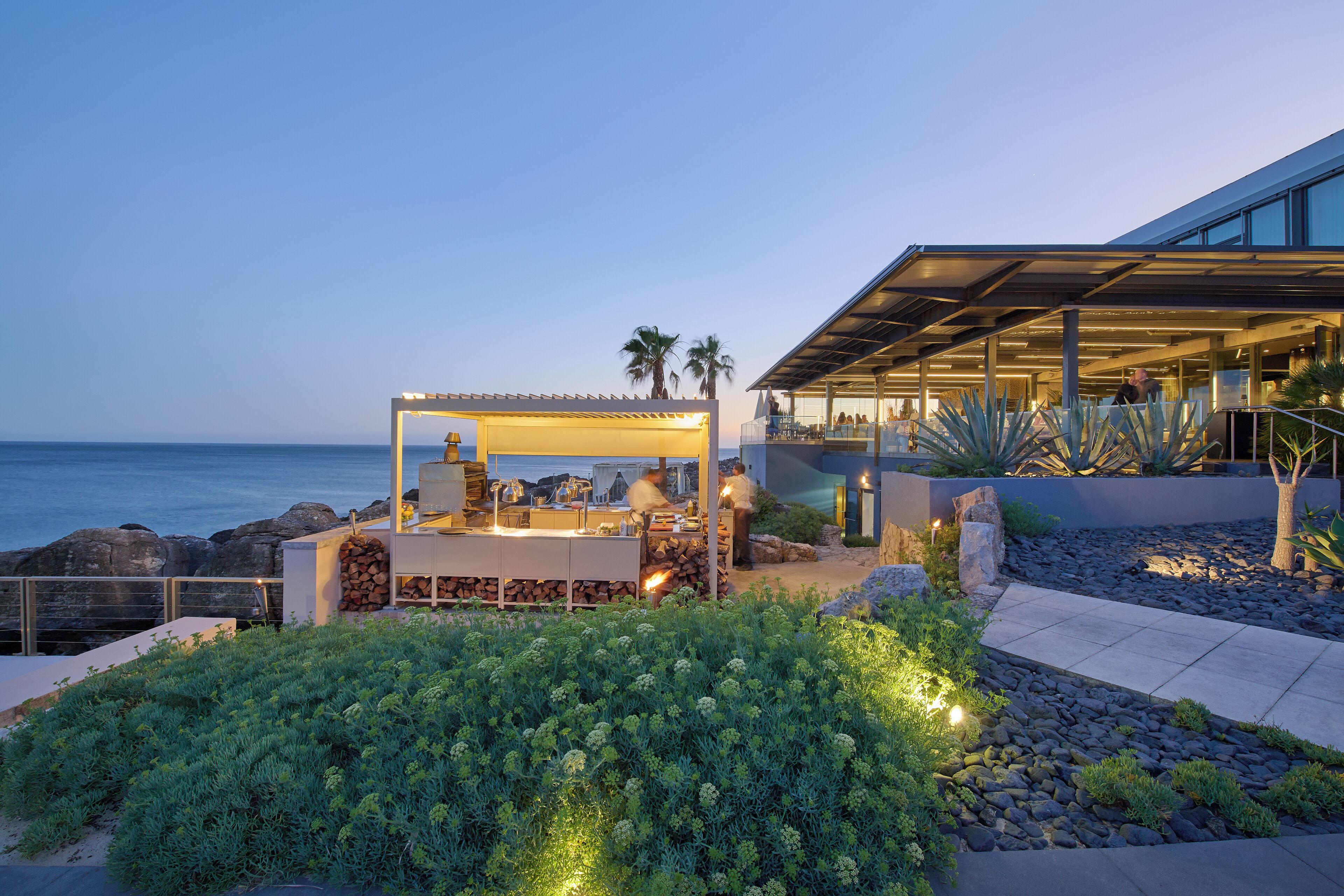 Um restaurante costeiro com área externa e mesas com vista para o mar, um edifício moderno com fachada de vidro e um jardim ajardinado.