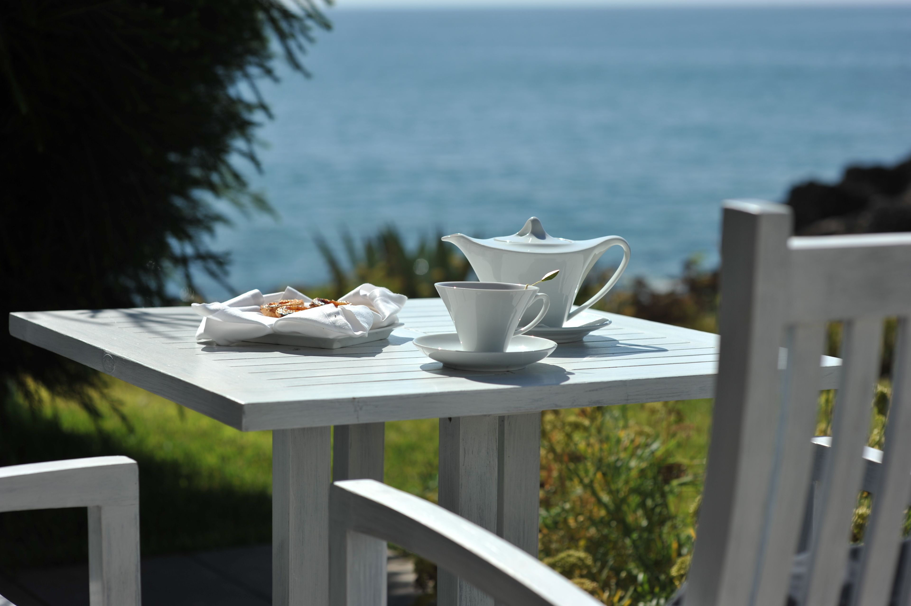 A white table with a teapot, cup, and saucer set on a wooden deck with a view of the ocean.