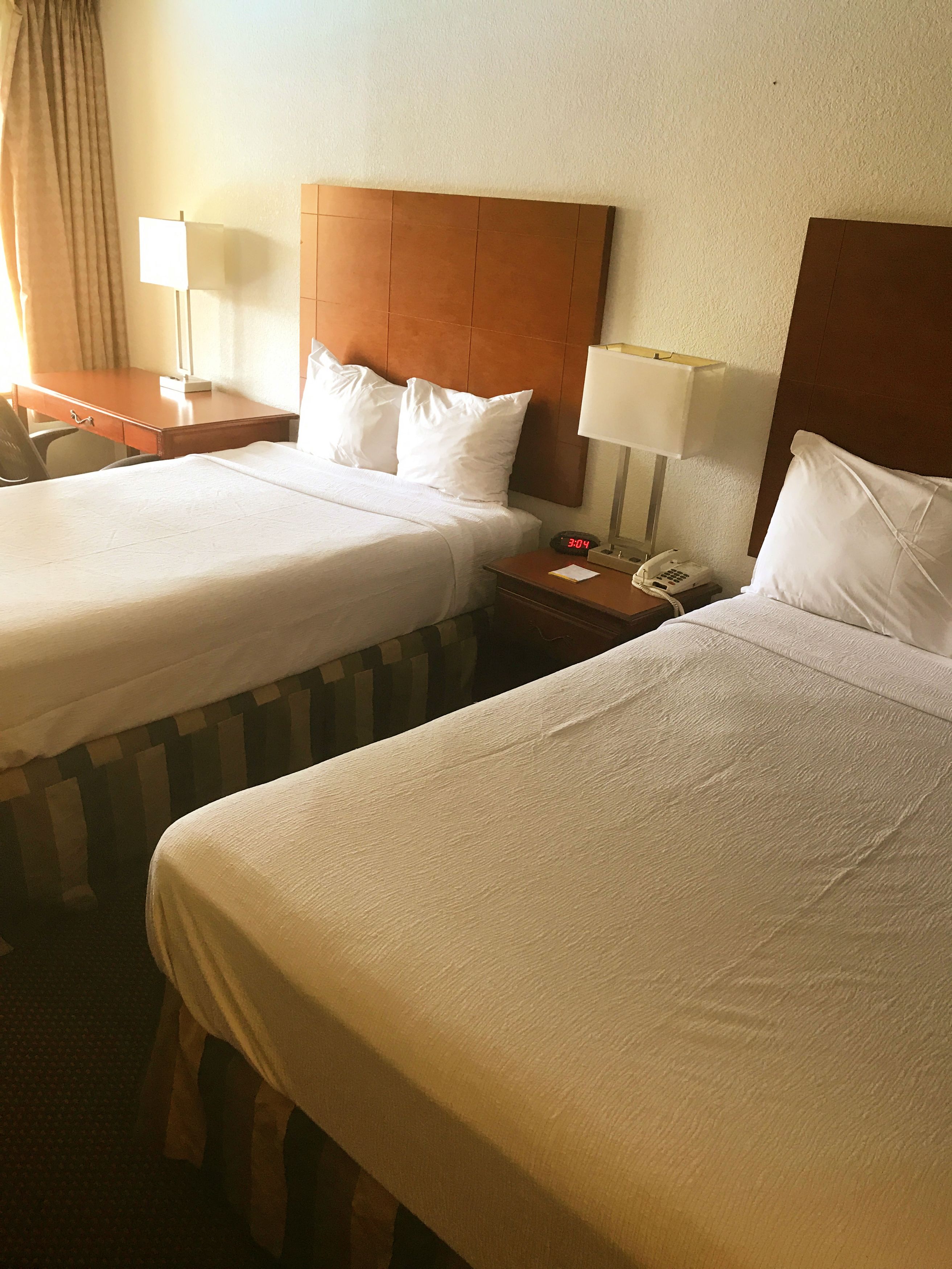 Una habitación de hotel con dos camas, cada una con ropa blanca y cabeceros de madera. Hay mesitas de noche con lámparas y un reloj que muestra la hora.