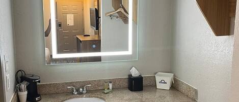 Un baño de hotel con un espejo grande, un lavamanos, dispensador de toallas y un cesto de basura.
