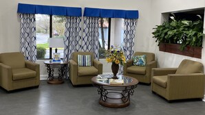 Un hall d’entrée avec quatre fauteuils beiges, une table basse en verre ornée d’un vase de fleurs et un lambrequin bleu et blanc à motifs.