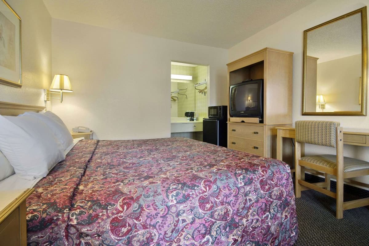 Una habitación de hotel con cama, televisión, escritorio, silla, espejo y kitchenette.