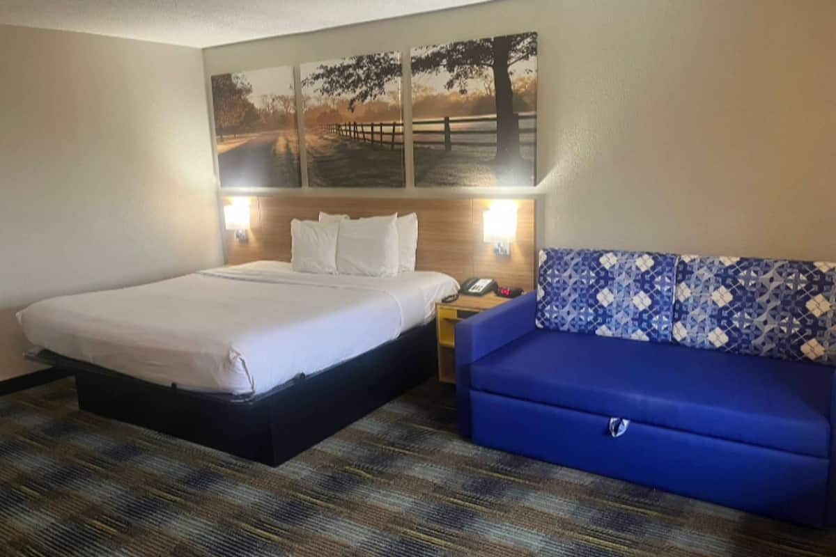Una habitación de hotel con una cama, un sofá azul, una mesita de noche con un teléfono y una obra de arte en la pared que muestra un sendero y árboles.