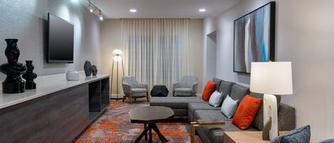 Een moderne hotelkamer met een grijze zithoek, een klein bijzettafeltje, een aan de muur bevestigde televisie en een groot raam met gordijnen.