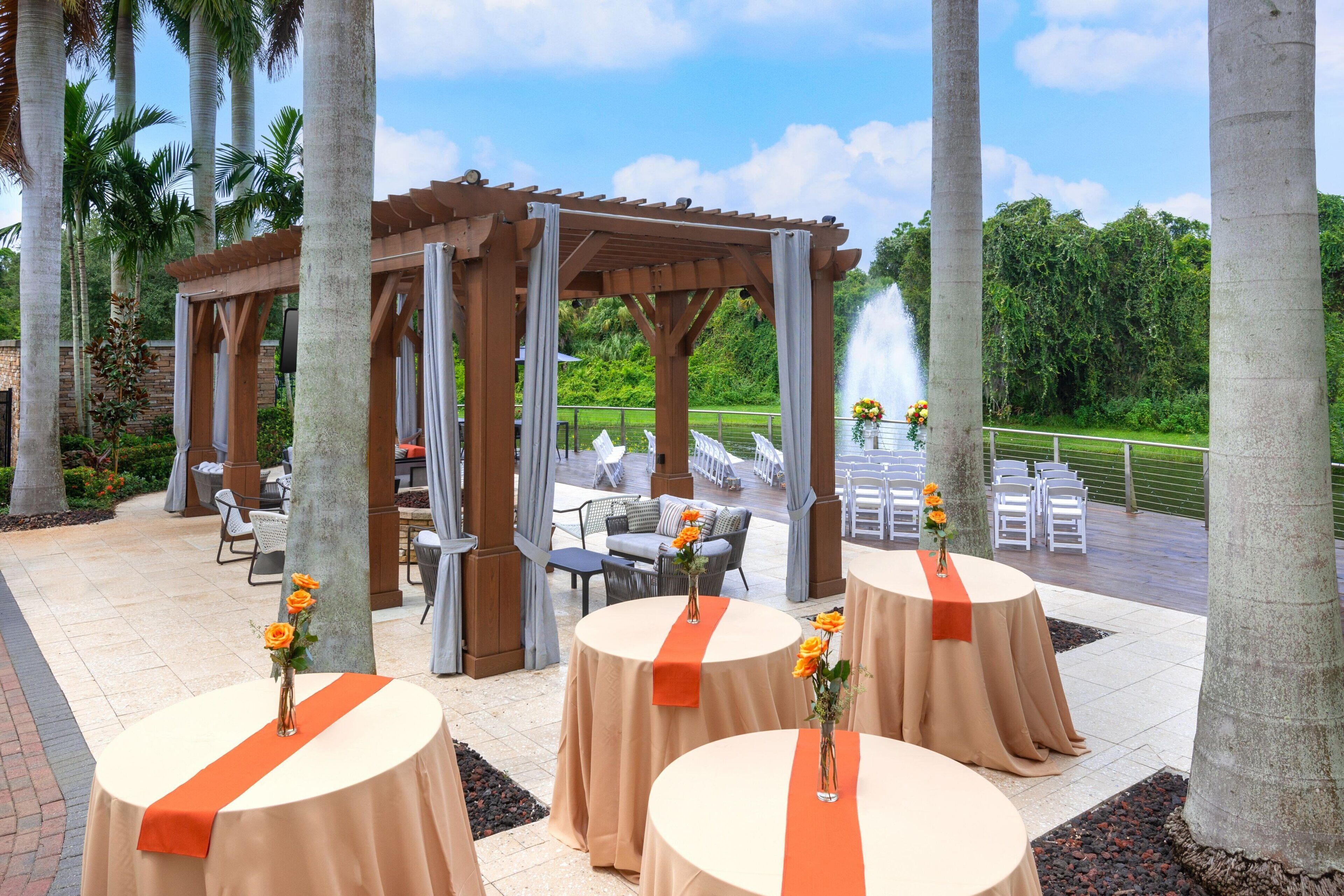 An outdoor patio area with round tables covered in beige tablecloths and orange sashes, a wooden pergola, and a fountain in the background.