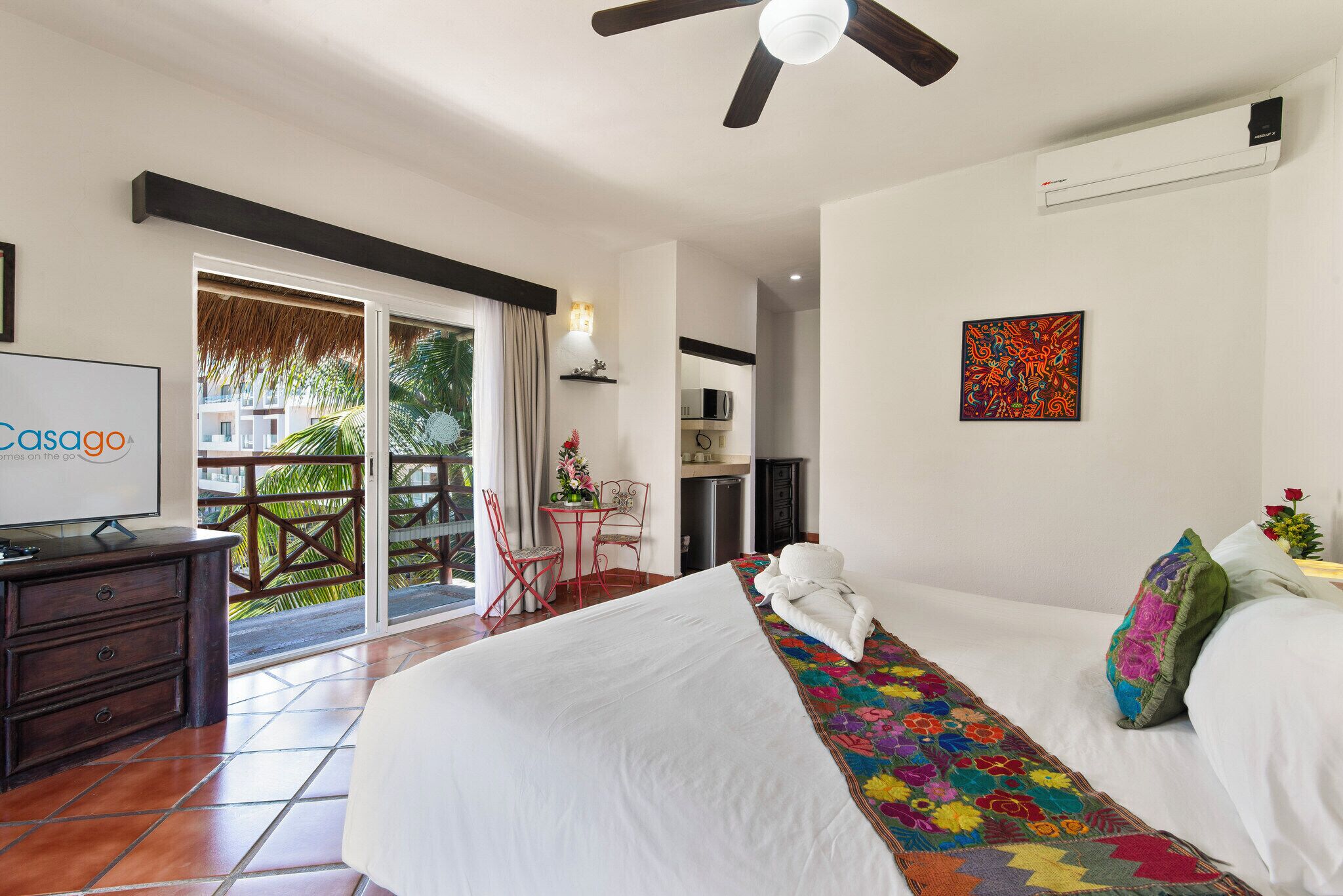 Habitación de hotel con cama, escritorio, silla, televisor y balcón con vista a palmeras.