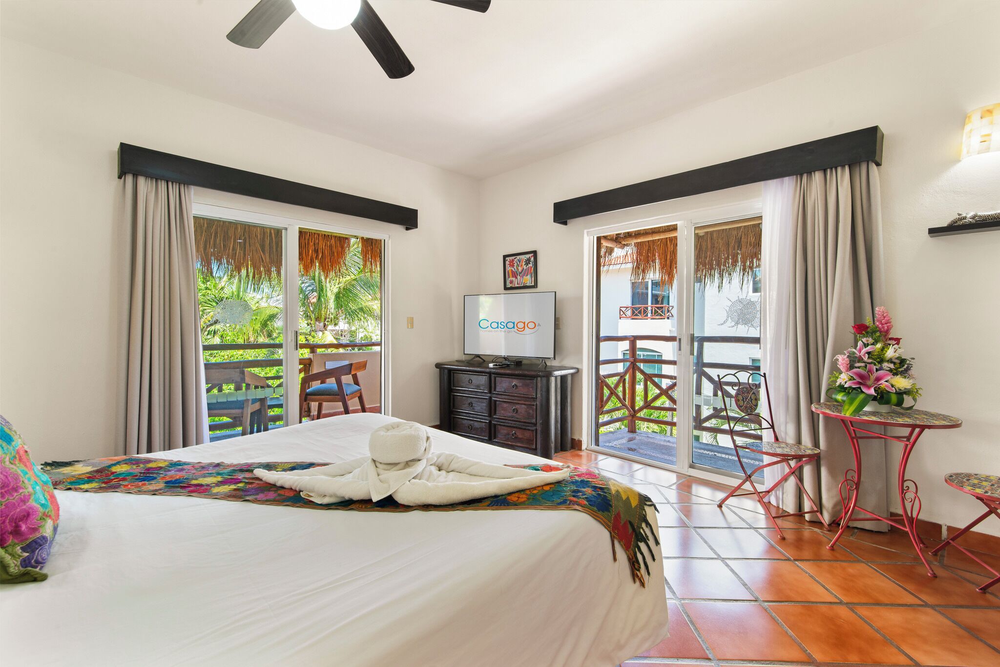 A hotel room with a bed, a TV, a balcony with a table and chairs, and a view of greenery outside.