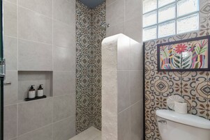 Un baño con baldosas estampadas, ducha, inodoro y repisa con artículos de aseo.
