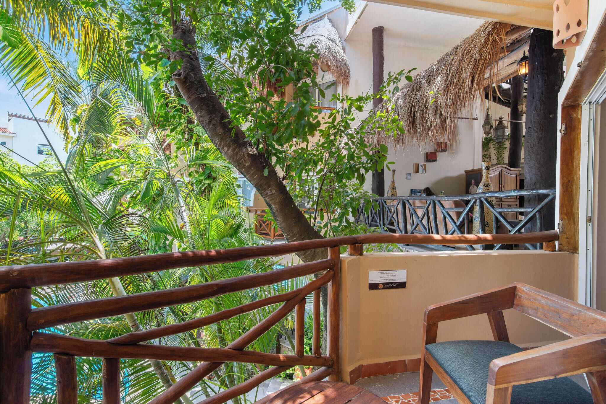 Una terraza de madera con techo de paja, rodeada de exuberante vegetación y vistas a un edificio con techo de paja y faroles colgantes.
