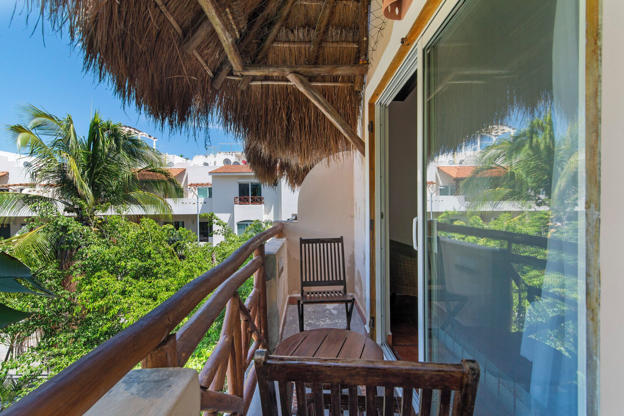 A balcony with a wooden railing, a thatched roof, and a view of a tropical landscape with palm trees and buildings.