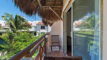 A balcony with a wooden railing, a thatched roof, and a view of a tropical landscape with palm trees and buildings.