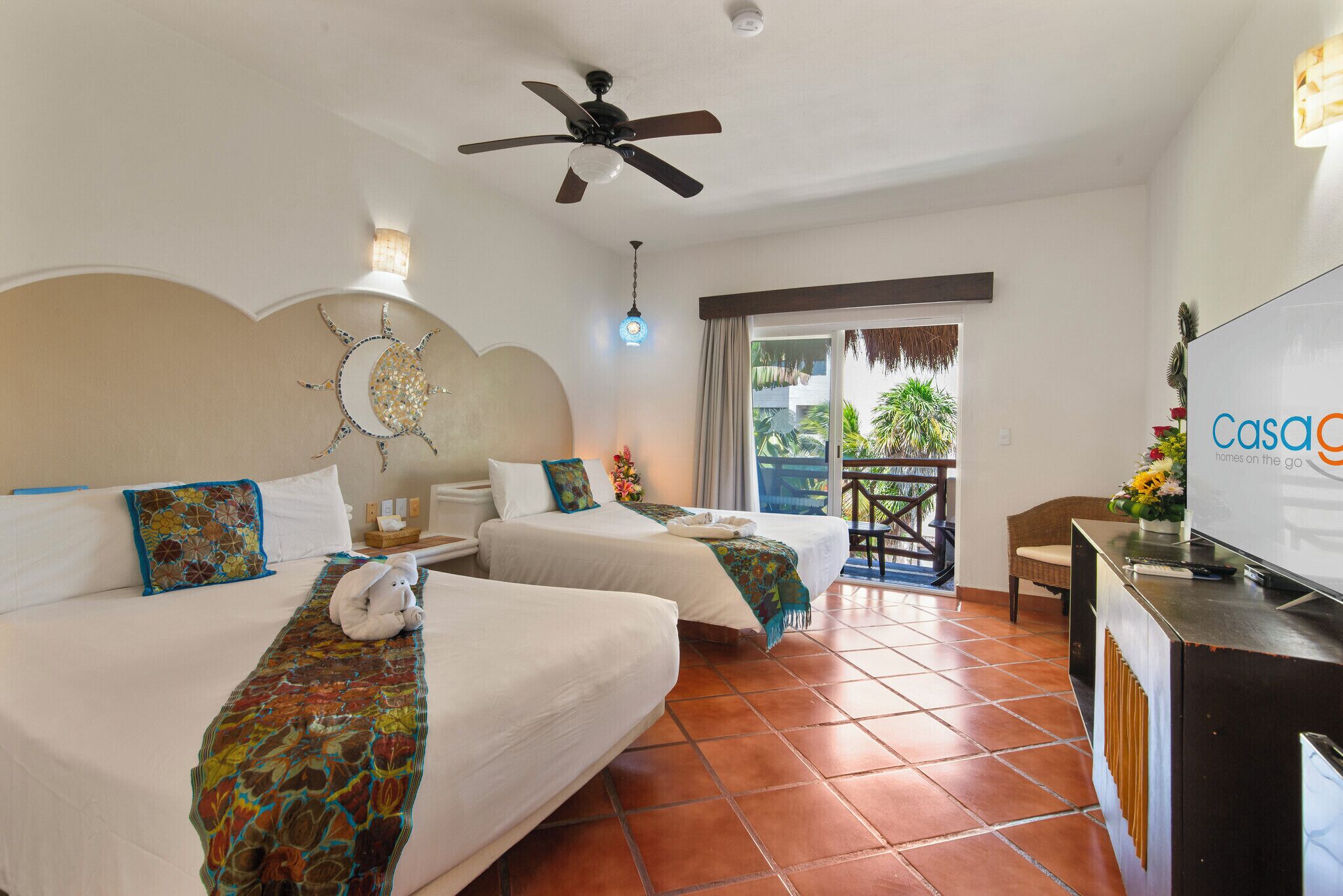 A hotel room with two beds, a ceiling fan, a TV, and a balcony with a view of palm trees.
