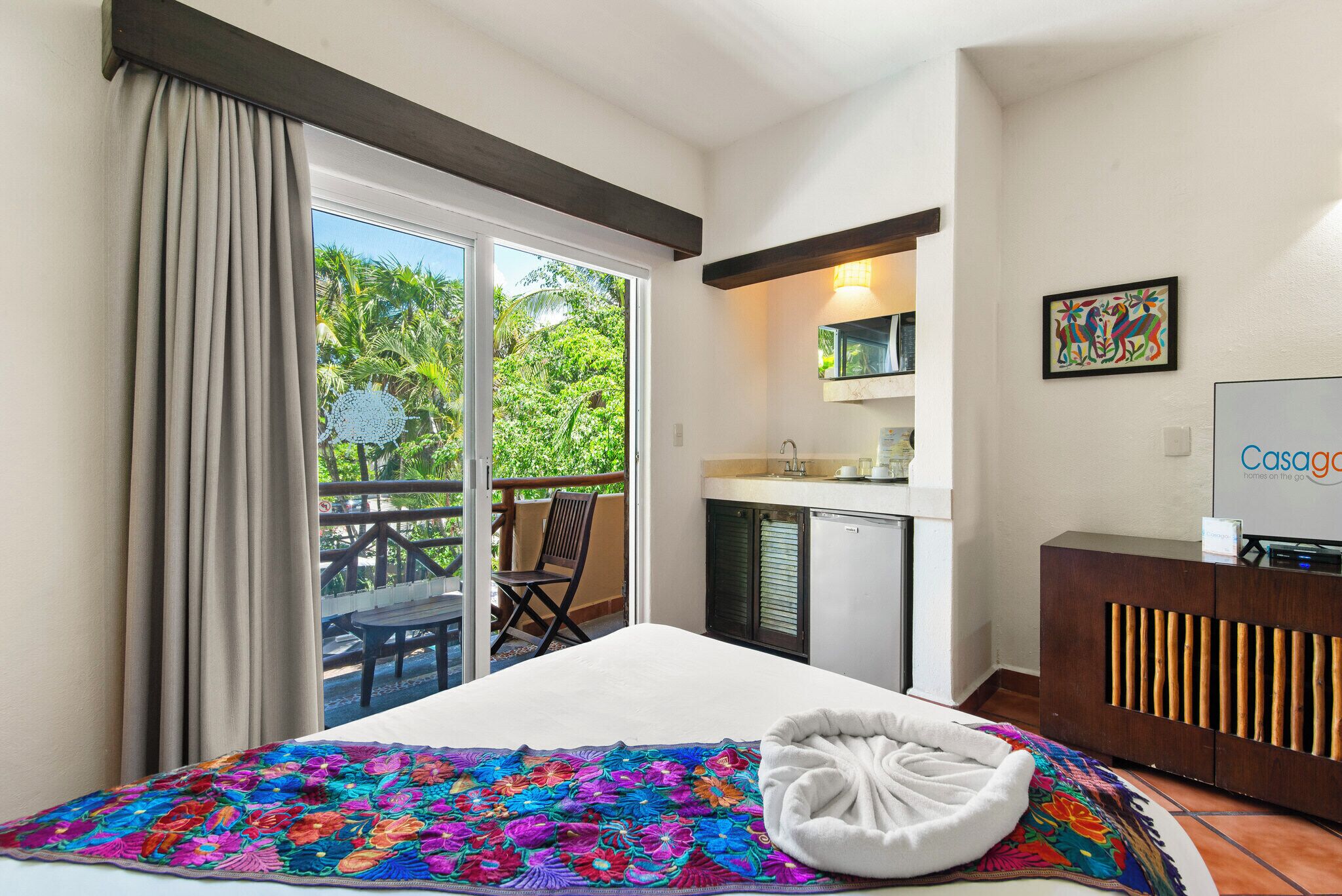 Une chambre d’hôtel avec un lit, une petite kitchenette, un balcon avec une table et des chaises, et une vue sur la verdure.