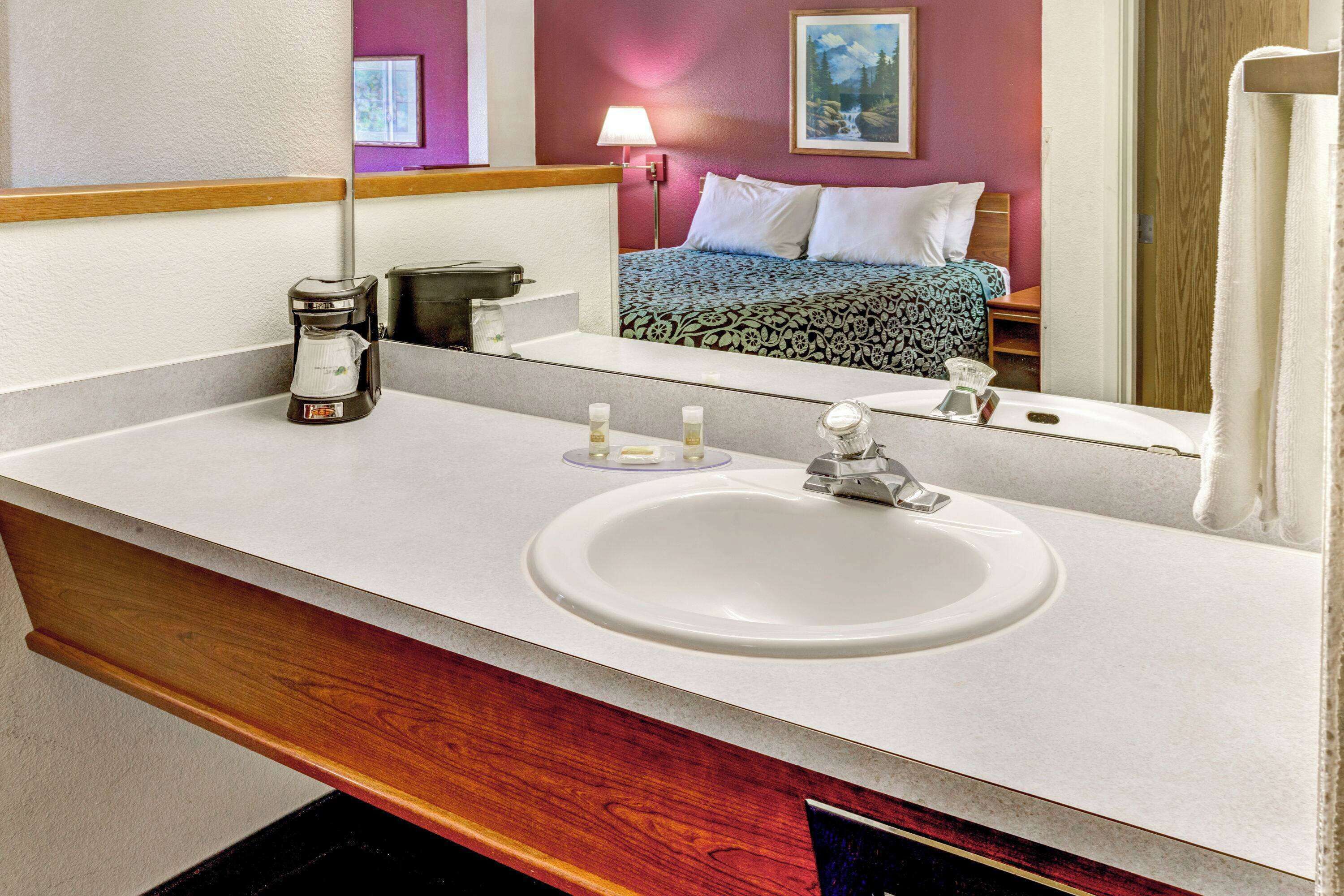 A hotel bathroom with a white sink, a coffee maker, and a mirror reflecting a bed and a painting on the wall.
