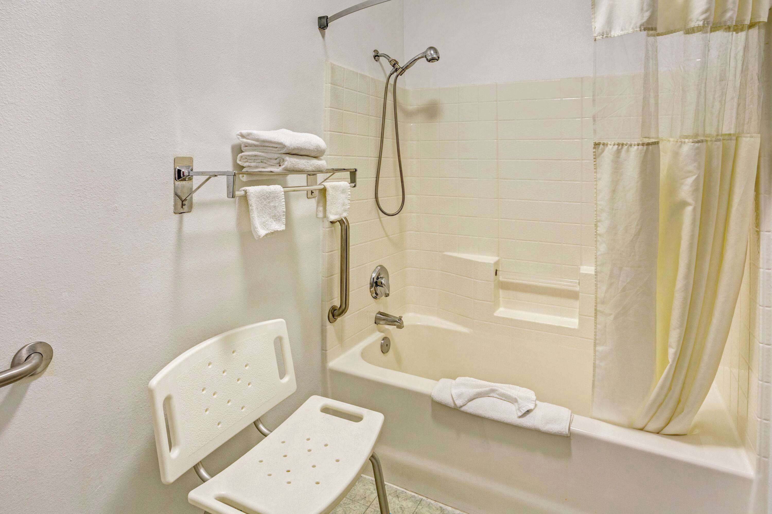 A bathroom with a white bathtub, a shower, a white grab bar, and a white chair.