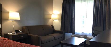 A hotel room with a bed, a desk with a lamp, a sofa, a small table, and a chair.