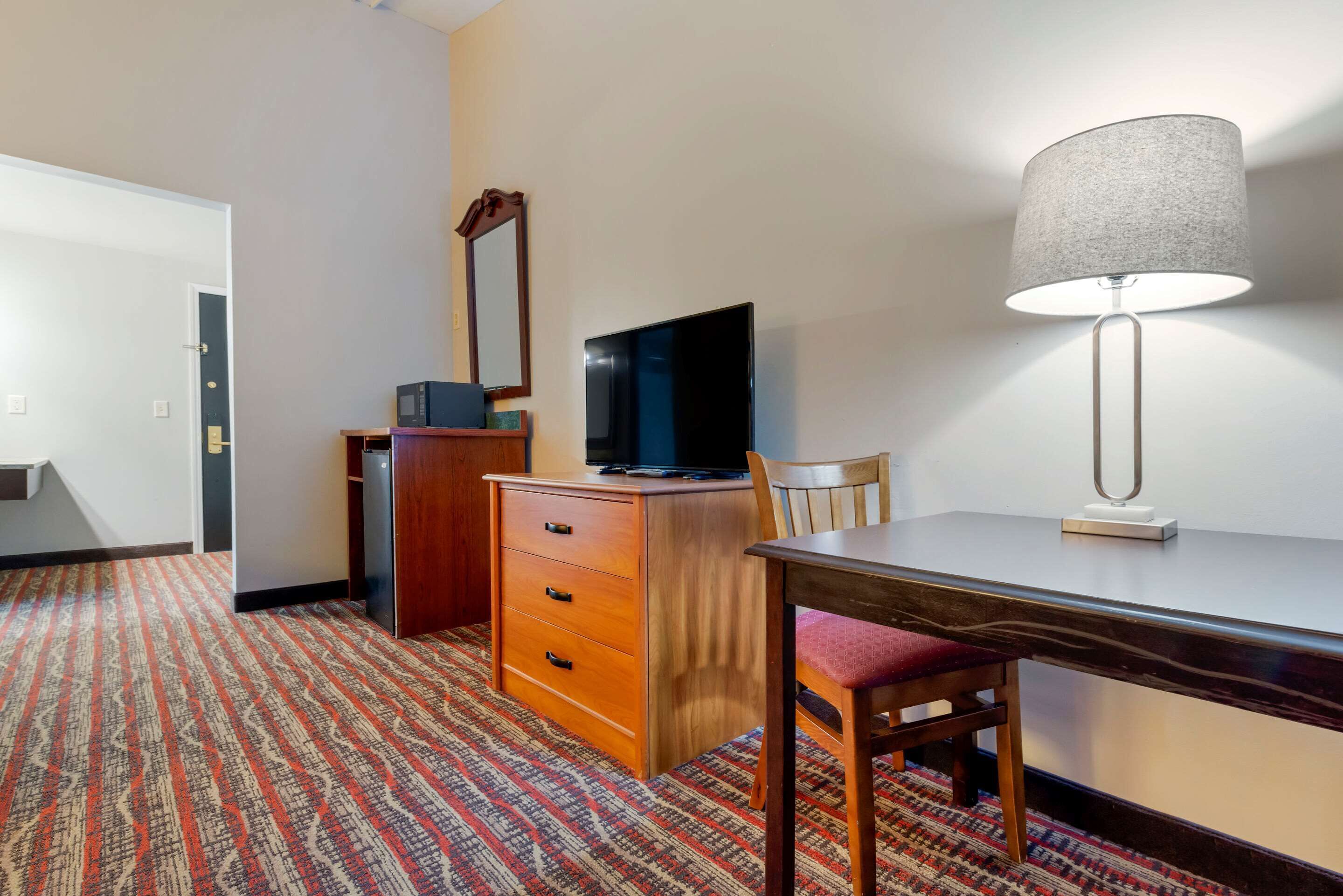 A hotel room with a wooden desk, a television, a lamp, a chair, and a patterned carpet.