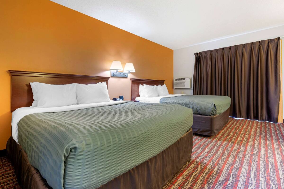 A hotel room with two beds, a headboard with lamps, an air conditioning unit, and a patterned carpet.