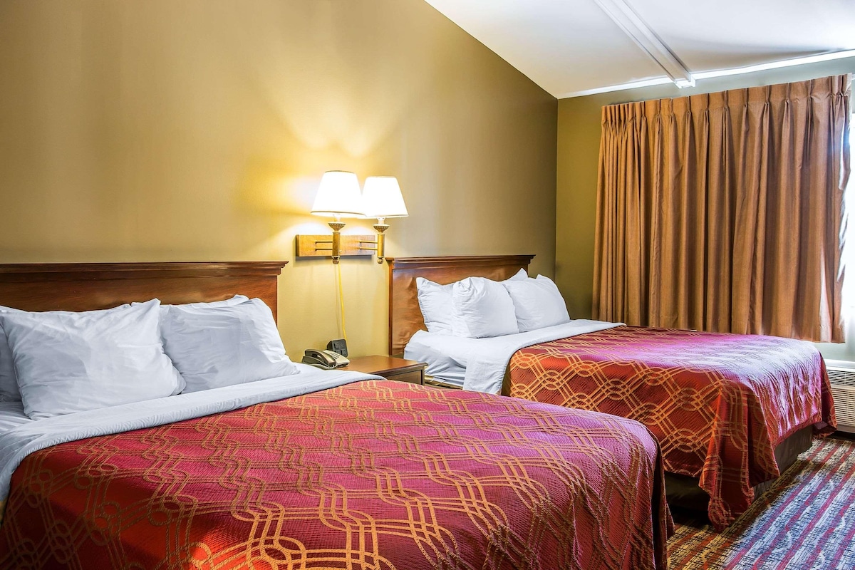A hotel room with two beds, each with white bedding and a patterned red blanket. There is a nightstand with a lamp and a phone. The room has a wooden headboard and a large window with curtains.