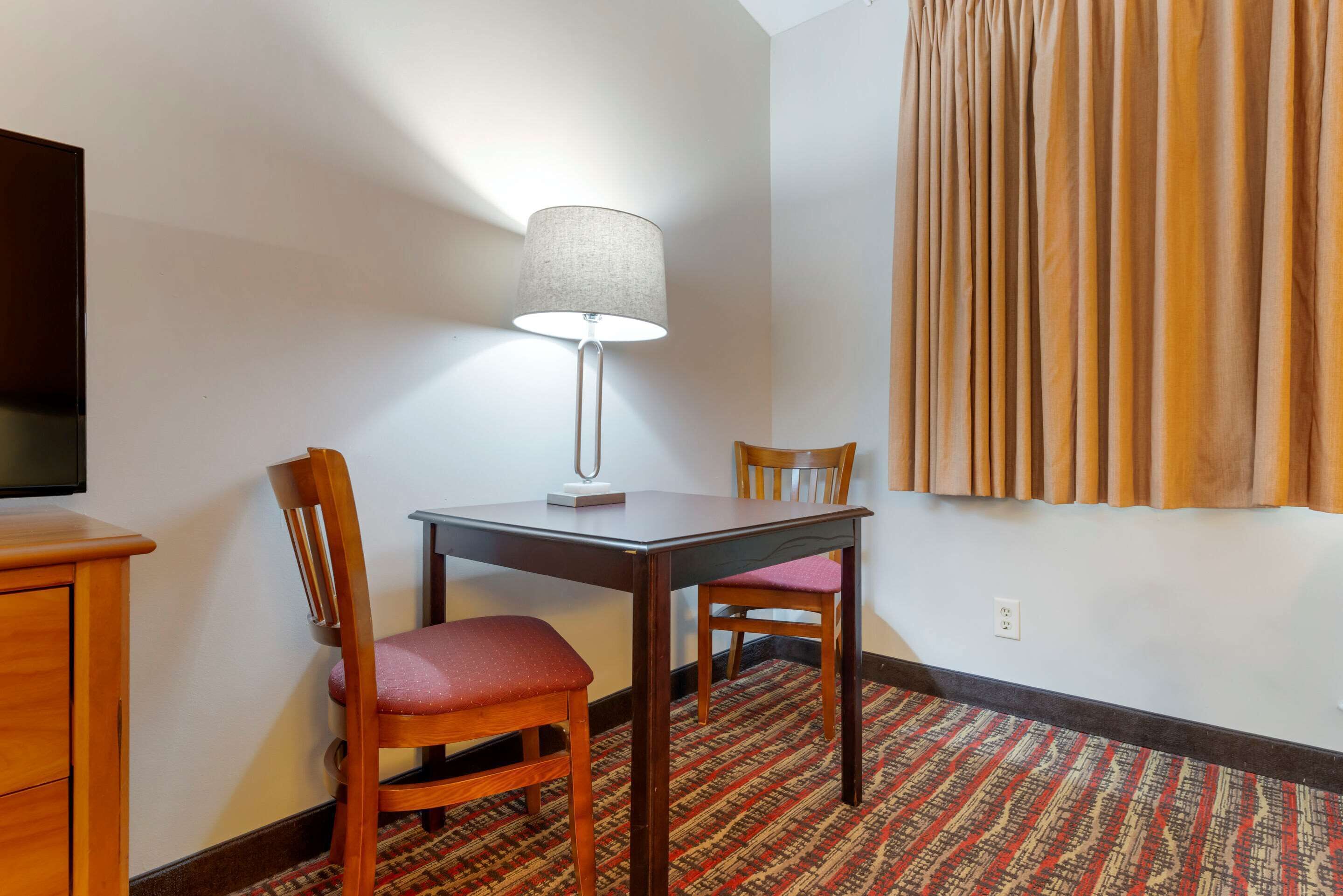 A hotel room with a wooden desk, two chairs, a lamp, a television, a patterned carpet, and a window with curtains.